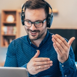 Ein Mann mit Brille und Kopfhörern spricht lächelnd in Videokonferenz vor dem Laptop.