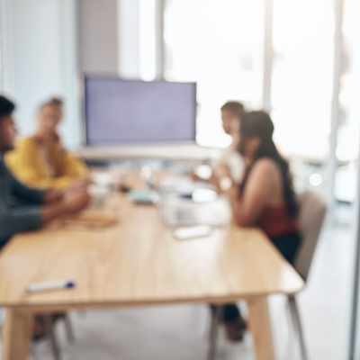 Mehrere Menschen sitzen an einem Besprechungstisch, die Darstellung ist verschwommen im Hintergrund ist ein großer Bildschirm zu sehen
