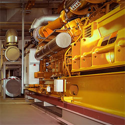 Das Bild zeigt eine industrielle Maschinenanlage in einer Produktionshalle mit einem großen gelben Generator oder Motor im Vordergrund.