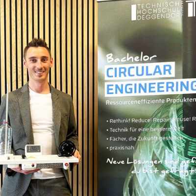 Ein lächelnder Bachelorabsolvent präsentiert stolz sein Projekt neben dem Informationsstand der Technischen Hochschule Deggendorf